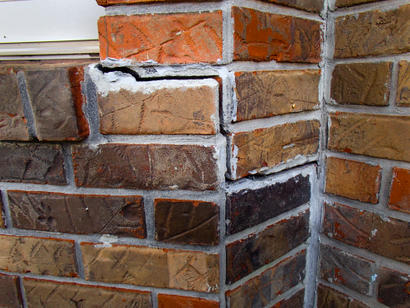 Cracked red and brown brick wall with visible mortar damage and repairs, located at a corner of a building.