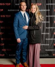 A couple poses on the red carpet; the man in a blue suit and the woman in a black jacket and long dress.