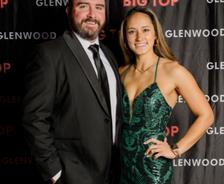 A man in a suit and a woman in a green dress pose together at an event with a "Glenwood Big Top" backdrop.