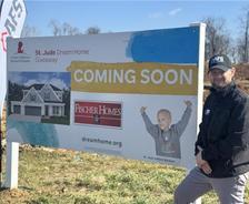 Man standing next to a "St. Jude Dream Home Giveaway Coming Soon" sign with a house and child pictured on it.