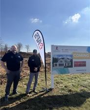 Two people stand beside a "St. Jude Dream Home" sign on a sunny day with a flag and clear sky in the background.