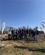 Group of people with shovels at a groundbreaking ceremony, clear sky, and tree line in the background.