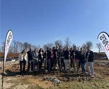 Group of people with shovels at a groundbreaking ceremony, clear sky, and tree line in the background.