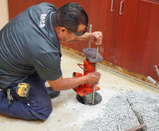 A worker installs a foundation pier in a room with wooden cabinets and debris on the floor.