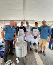 Six men standing under a tent, some wearing aprons, posing beside a large pot on a burner.