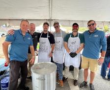 Six men standing under a tent, some wearing aprons, posing beside a large pot on a burner.