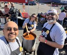 Three people smiling at a chili cook-off, wearing aprons and sunglasses under a sunny sky.