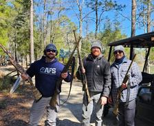 Three people holding rifles stand outdoors near a forest and a utility vehicle, all wearing casual clothing and hats.