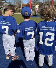 Three kids in blue baseball jerseys with numbers 7, 12, and 3, standing on a playground with a rubber surface.