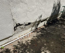 Cracked exterior wall with exposed damage near piping, surrounded by shadows and debris on the ground.