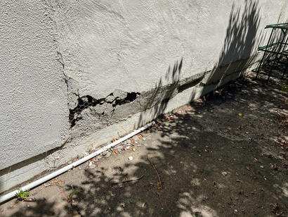 Cracked exterior wall with exposed damage near piping, surrounded by shadows and debris on the ground.