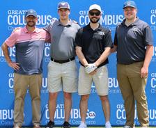 Four men in golf attire pose together in front of a blue "Greater Columbus" event backdrop outdoors.