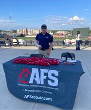 Man stands behind an AFS company booth at a stadium, with promo items on the table and a cityscape behind.