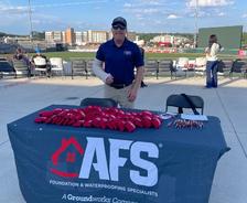 Man stands behind an AFS company booth at a stadium, with promo items on the table and a cityscape behind.