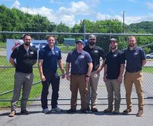Six men standing outside in front of a chain-link fence on a sunny day, posing for a group photo.
