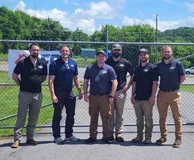 Six men standing outside in front of a chain-link fence on a sunny day, posing for a group photo.