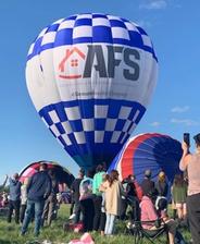 Crowd watching a large blue and white hot air balloon with "AFS" logo at a festival under a clear sky.