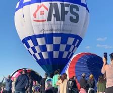 Crowd watching a large blue and white hot air balloon with "AFS" logo at a festival under a clear sky.