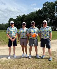 Four men wearing matching colorful shirts and sunglasses stand outside on a sunny day near trees and grass.