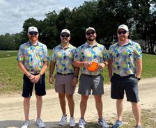 Four men wearing matching colorful shirts and sunglasses stand outside on a sunny day near trees and grass.