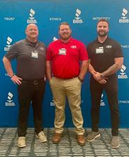 Three men pose and smile in front of a blue BBB backdrop with words like "trust," "ethics," and "integrity.
