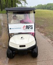 Two people sit in a golf cart on a rainy path, with sponsor signs on the windshield.