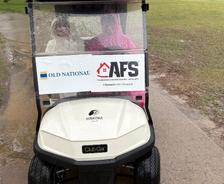 Two people sit in a golf cart on a rainy path, with sponsor signs on the windshield.