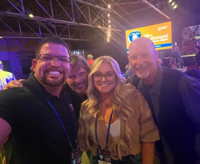 Four smiling people pose for a selfie at Songwriters Night, an AFS event, with Annual Sponsors featured on the large screen behind them.
