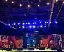 Concert stage set for Songwriters Night by AFS, featuring guitars, stools, and colorful lights in the background.