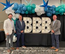 Three men in suits stand before large, lit "BBB" letters and balloons at the BBB Torch Awards, celebrating ethics in business.