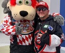 A person in sports gear poses with the Huntsville Havoc’s large, gray wolf mascot in a red and white jersey.