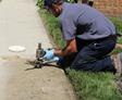Person in work attire and gloves kneeling on a sidewalk, inspecting or repairing a plumbing fixture near grass.