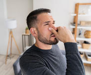Man in a gray sweater pinching his nose, appearing bothered, standing in a living room with shelves in the background.