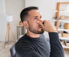 Man in a gray sweater pinching his nose, appearing bothered, standing in a living room with shelves in the background.