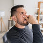 Man in a gray sweater pinching his nose, appearing bothered, standing in a living room with shelves in the background.