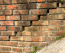 Old brick wall with loose bricks and visible weathering, next to a sloped concrete surface with some greenery.