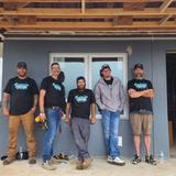 Five smiling men stand in front of a house under construction, wearing matching shirts and caps.