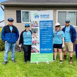 Four people in volunteer shirts, hard hats, and jeans stand by a Habitat for Humanity banner outside a house.