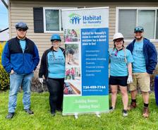 Four people in volunteer shirts, hard hats, and jeans stand by a Habitat for Humanity banner outside a house.