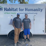 Two people smiling in front of a Habitat for Humanity of Kent County trailer on a sunny day.