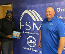 Two men smiling and holding a certificate, standing in front of a "FSM Grand Rapids" sign.