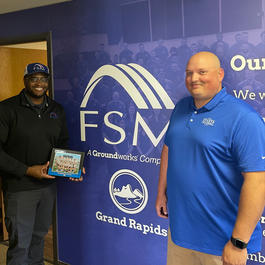 Two men smiling and holding a certificate, standing in front of a "FSM Grand Rapids" sign.