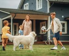 A family with two kids and a dog play soccer together in the yard in front of a house.