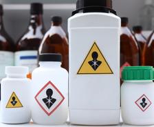 Four chemical bottles with hazard symbols indicating toxicity, with brown bottles blurred in the background.
