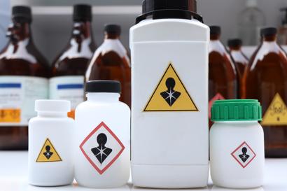 Four chemical bottles with hazard symbols indicating toxicity, with brown bottles blurred in the background.