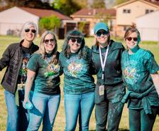 Five smiling women with sunglasses stand together outdoors, covered in blue powder, wearing matching event T-shirts.