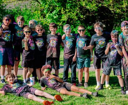 A group of kids with colorful powder on their clothes and faces smile together outside on a sunny day.