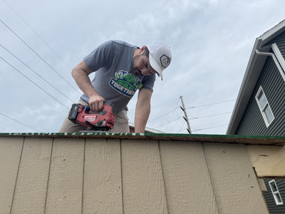 Man using a power tool to work on the roof of a wooden structure under a cloudy sky.