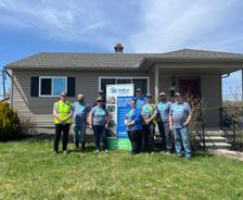 A group of people stand in front of a house and a Habitat for Humanity sign on a sunny day.
