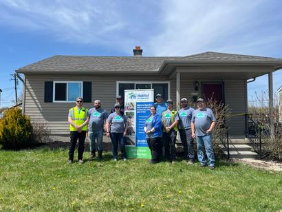 A group of people stand in front of a house and a Habitat for Humanity sign on a sunny day.